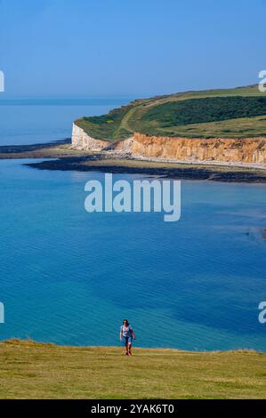 Vista prospettica di una donna che cammina verso una ripida collina erbosa su Seven Sisters con uno sfondo del mare e Seaford Head. East Sussex, Inghilterra. Foto Stock