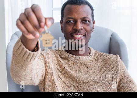 L'uomo afroamericano mostra felicemente le chiavi della sua nuova casa mentre è seduto su una poltrona, illuminata dalla luce naturale da una finestra Foto Stock