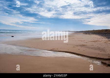 Una grande distesa di spiaggia sabbiosa che si estende all'orizzonte sotto un cielo nuvoloso blu. Un flusso d'acqua attraversa la sabbia e scorre verso il mare Foto Stock