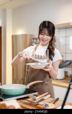 Una giovane donna asiatica affascinante e positiva in un grembiule sta cucinando in cucina, afferrando salsicce da una padella con pinze e mettendole su un piatto. hom Foto Stock