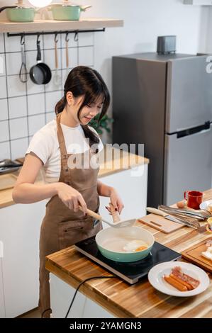 Una giovane donna asiatica bella e felice in grembiule sta cucinando in cucina, friggendo un uovo in una padella, preparando un uovo con il lato soleggiato per la sua colazione. cavolo Foto Stock