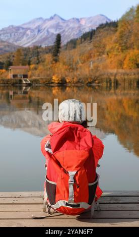 La ragazza turistica con lo zaino sulla schiena ammira una fantastica vista delle montagne sedute sulla riva del lago Foto Stock