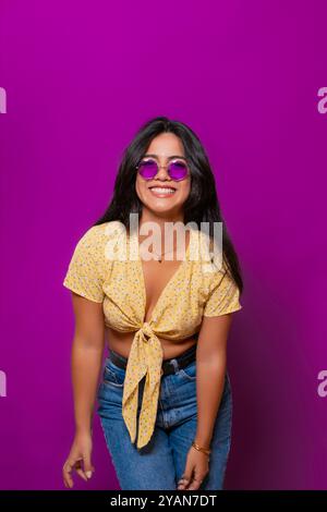 Donna allegra che indossa un top floreale giallo e occhiali da sole viola, sorridente mentre si piega in avanti in una posa giocosa su uno sfondo viola. Foto Stock