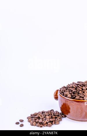 Una ciotola marrone calda piena di chicchi di caffè naturali, con pochi chicchi sparsi intorno, il tutto su uno sfondo bianco brillante Foto Stock
