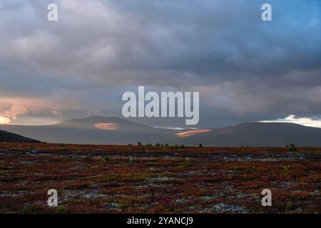 Nuvole spettacolari gettano ombre sul paesaggio, mentre il sole basso illumina le montagne lontane e la colorata vegetazione della tundra Foto Stock