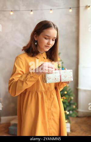 Una giovane donna sta aprendo con gioia un regalo di Natale, indossando un abito giallo brillante che irradia felicità. Ragazza che tiene in mano una confezione regalo per la vigilia di Capodanno Foto Stock