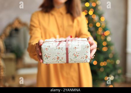 Una giovane donna sta aprendo con gioia un regalo di Natale, indossando un abito giallo brillante che irradia felicità. Ragazza che tiene in mano una confezione regalo per la vigilia di Capodanno Foto Stock