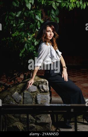 Una giovane donna che indossa un abito alla moda in stile retrò posa elegantemente su gradini di pietra, incorniciati da lussureggianti fogliame verde, in un ambiente all'aperto sereno duri Foto Stock