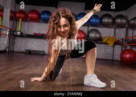 Una donna dai capelli lunghi e ricci danzerà dinamicamente in una sala fitness piena di attrezzature per l'esercizio fisico Foto Stock