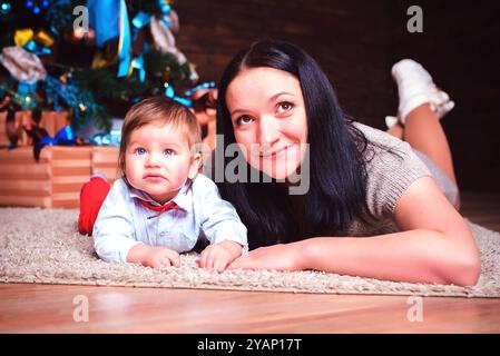 Una madre allegra e allegra gioca con amore con il suo adorabile bambino proprio accanto al bellissimo albero di Natale Foto Stock