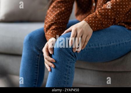Donna asiatica seduta sul divano che tiene il ginocchio, indossa jeans e pois top. L'immagine cattura il concetto di dolore articolare, salute e relax a casa. Attenzione al disagio e al benessere. Foto Stock
