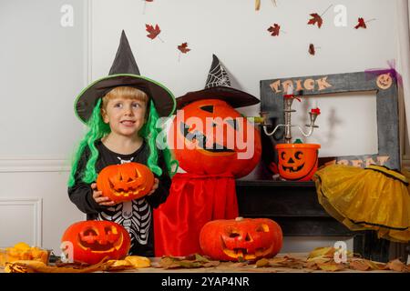 Un bambino con una parrucca verde, il cappello della strega festeggia Halloween, circondato da zucche intagliate e foglie autunnali Foto Stock