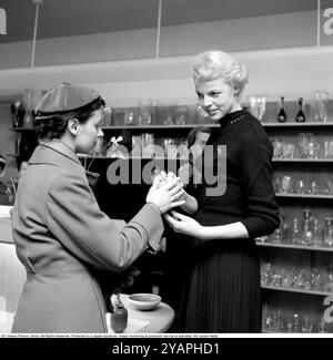 Shopping negli anni '1950 Una giovane donna in un negozio in cui una donna di vendita presenta alcuni dei negozi della selezione di ceramiche pregiate e oggetti decorativi progettati dal famoso designer Stig Lindberg. Si vede che tiene in mano un vaso che lo mostra correttamente per la cliente donna. 1956 Conard rif. 3181 Foto Stock