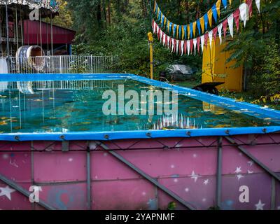 Piscina vuota e tranquilla per i giocattoli galleggianti con foglie cadute sulla superficie dell'acqua e un'alta giostra a due piani chiusa per la stagione fredda. Foto Stock