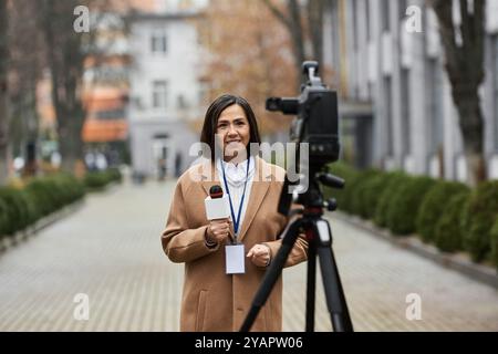 Un giornalista fiducioso si trova all'aperto in un cappotto beige, tenendo un microfono e preparandosi a segnalare le notizie. Foto Stock