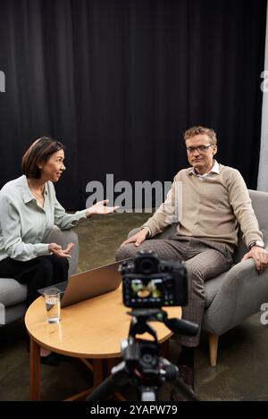 Una giornalista di mezza età conduce un'intervista professionale con un uomo seduto su un elegante divano. Foto Stock