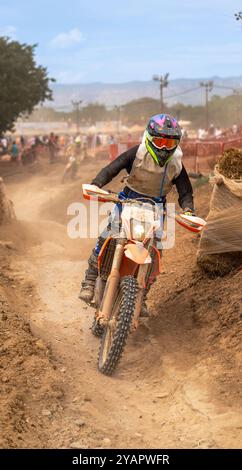 Pilota di motocross coperto di fango e polvere che salta la sua moto al cambio di pendenza su una pista sterrata durante una gara di resistenza di motocross con spettatori. Foto Stock