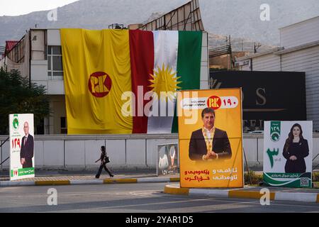 Duhok, Iraq. 13 ottobre 2024. Una giovane donna oltrepassa gli striscioni elettorali dei candidati del partito dell'Unione Patriottica del Kurdistan (PUK) e l'immagine del Partito Democratico del Kurdistan (KDP/PDK) del vicepresidente del partito Nechirvan Barzani in vista delle elezioni del Parlamento regionale del Kurdistan nella città di Duhok, Iraq. (Foto di Ismael Adnan/SOPA Images/Sipa USA) credito: SIPA USA/Alamy Live News Foto Stock