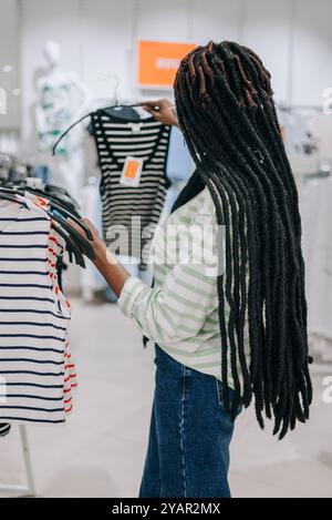 Giovane donna afro-americana elegante con dreadlock che sfoggiano in boutique di moda alla moda, selezionando un'elegante camicia a righe bianche e nere da un appendiabiti con particolare attenzione alle opzioni di prezzo in vendita Foto Stock