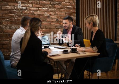 Quattro professionisti si impegnano attivamente in una discussione attorno a un tavolo in un ufficio elegante con pareti in mattoni a vista. Sembrano concentrati e animati Foto Stock