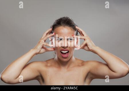 Foto che cattura l'intensa espressione di una giovane donna in difficoltà con le mani sulla testa, esprimendo frustrazione o stress su uno sfondo semplice. Foto Stock