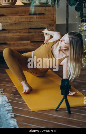 Una donna che indossa un top marrone e pantaloni si sta esercitando con una posa da yoga su un tappetino giallo, tenendo il piede con la mano. Un treppiede è posizionato nelle vicinanze, a indicare che è in corso un video Foto Stock