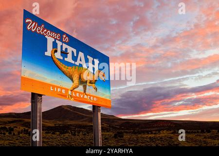 Un cartello "benvenuto nello Utah" con un dinosauro si erge lungo una strada rurale dello Utah con un tramonto sullo sfondo. Foto Stock