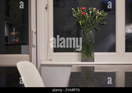 fiori nuovi tagliati in un vaso di vetro e un vaso di vetro vuoto su un ripiano di fronte alla finestra di notte con un leggero riflesso della zona cucina Foto Stock