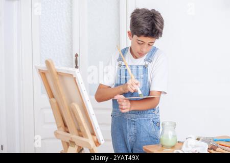 Un ragazzo sta dipingendo un quadro su una tela. Indossa una tuta blu e una camicia bianca. Il dipinto è in corso e il ragazzo sta usando un pennello Foto Stock