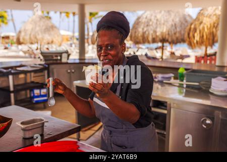 Donna afro americana che serve gelato al ristorante all'aperto sulla spiaggia con sfondo tropicale. Foto Stock