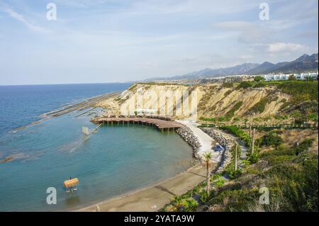 Il tranquillo litorale si dispiega, rivelando un rustico molo in legno che si estende nel mare calmo, incorniciato da una vegetazione lussureggiante e da colline lontane sotto morbide morni Foto Stock