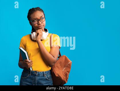 Pensieroso ragazza nera della studentessa che pensa in piedi su sfondo di studio blu Foto Stock