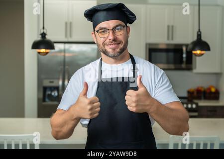 Cuoco felice con il pollice in su. Ispanico in uniforme baker. Cucina e cucina. Chef maschile in uniforme da lavoro, grembiule nero, cappello da chef. Foto Stock