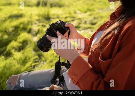 Una donna siede sull'erba, tenendo in mano una macchina fotografica mentre esamina le sue foto. La scena viene catturata all'aperto in una giornata di sole, concentrandosi su fotogr Foto Stock