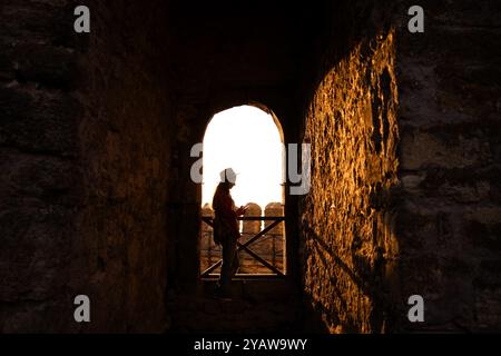Il viaggiatore si erge come una silhouette contro il bagliore dorato del tramonto, incorniciato dall'arco di pietra di un'antica fortezza. Le ombre e la luce creat Foto Stock