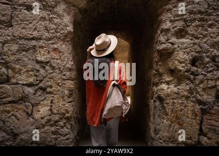 Un viaggiatore che indossa un cappello e porta con sé uno zaino cammina attraverso uno stretto passaggio in pietra all'interno di un'antica fortezza. struttura dei muri in pietra con agenti atmosferici Foto Stock