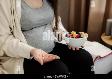 Primo piano di una giovane donna incinta che tiene in mano una ciotola di frutta e pillola, vitamine, integratori. Gravidanza alimentazione sana, mangiare. Fare una scelta. Concetto medico, farmaceutico e sanitario Foto Stock