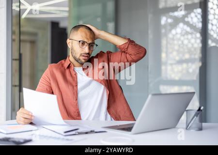 Uomo preoccupato in ufficio che tiene un documento mentre guarda un notebook con un'espressione stressata. L'ambiente professionale suggerisce preoccupazioni aziendali, stress e scenari decisionali. Foto Stock