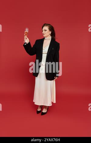 Una giovane donna vestita elegantemente con un blazer nero e un lungo abito bianco solleva un bicchiere di champagne. Foto Stock
