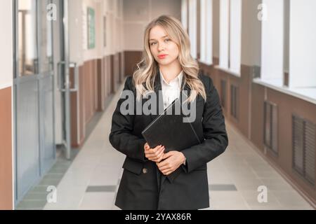 Bella donna bionda con un abito da lavoro nero e una camicia bianca con una cartella in un ufficio moderno. Donna d'affari di successo Foto Stock