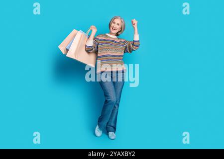 Foto a tutta lunghezza dell'estatico pensionato indossa jeans pullover ornamentali che tengono le borse della spesa isolate su sfondo blu Foto Stock