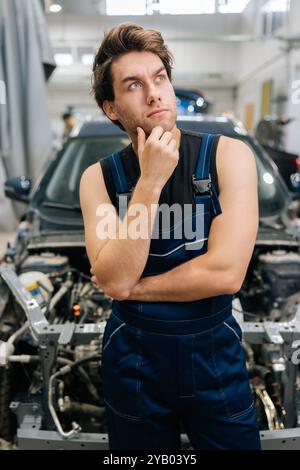 Ritratto verticale di un riparatore confuso che guarda lontano in un'officina auto in piedi in auto in riparazione. Un meccanico dubbioso che cerca di risolvere il problema in auto Foto Stock