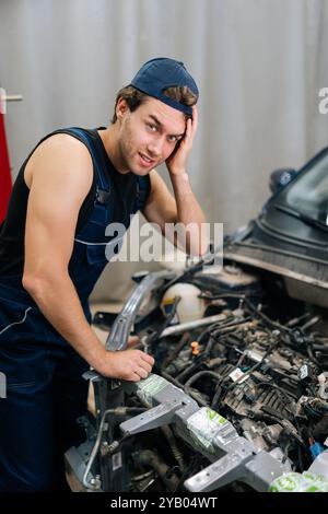 Il ritratto verticale di un giovane meccanico frustrato sembra preoccupato ed esasperato mentre lavora per riparare il motore di un'auto in garage, utilizzando gli attrezzi per Foto Stock
