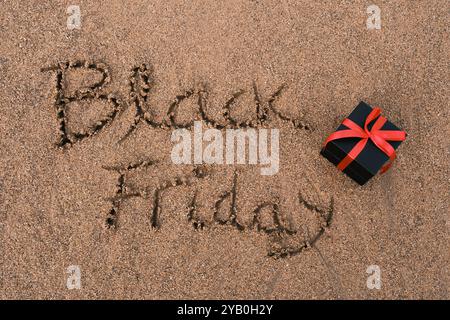 Testo del Black Friday scritto in sabbia con confezione regalo nera e nastro rosso Foto Stock