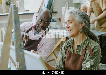 Colpo ad angolo alto di marito afroamericano che punta su tela mentre dà consigli alla sua amata moglie caucasica Foto Stock