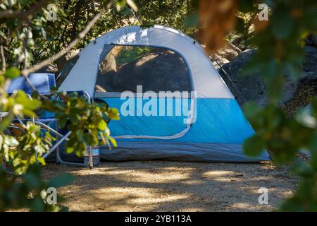 Una tenda blu e grigia si trova in un'area boscosa. Due sedie siedono di fronte alla tenda e una bottiglia d'acqua poggia su una delle sedie. Foto Stock