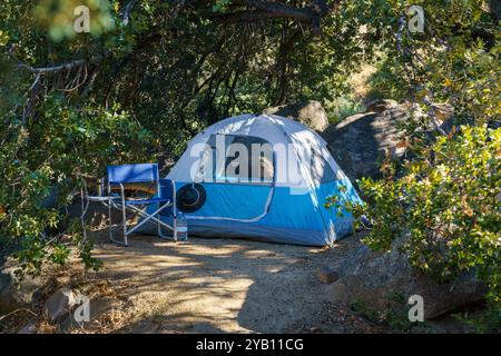 Una tenda blu e grigia si trova in un'area boscosa. Due sedie siedono di fronte alla tenda e una bottiglia d'acqua poggia su una delle sedie. Foto Stock