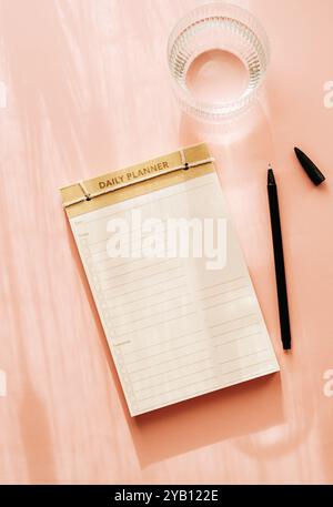 Pianificatore quotidiano minimalista piatto dal design semplice, con caselle di controllo e linee per le attività. Superficie rosa accanto a una tazza di vetro, creando una a pulita Foto Stock