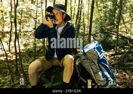 Uomo anziano seduto su un ceppo d'albero e che fa fotografie con una macchina fotografica. Uomo con fotocamera retrò e zaino che fa foto. Uomo anziano seduto su tre Foto Stock