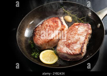 Due bistecche di lombo di maiale sfrigolanti e cotte a vapore in una padella calda di ghisa con erbe aromatiche e fette di limone, cucinare un pasto con carne arrosto, copiare lo spazio Foto Stock